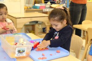 A young girl uses a stamp to press shapes into orange playdough on a tray, with a bin labeled 'Playdoh Tools' nearby in a classroom art area.